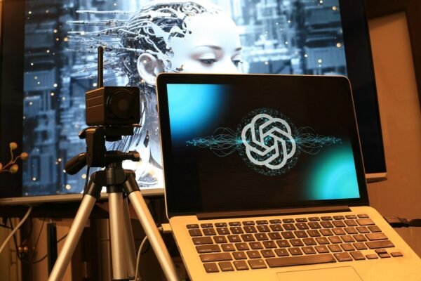 A laptop with an OpenAI logo connected to a camera on a tripod in the background is a screen with a robot photo